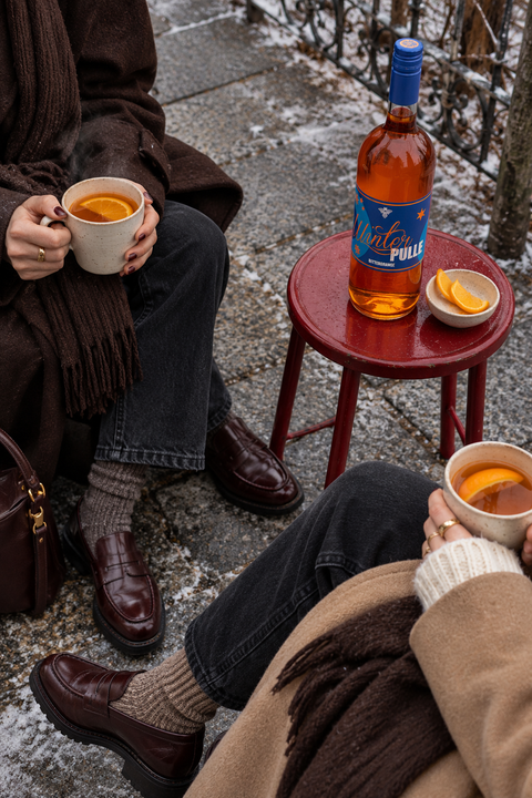 WinterPulle Bitter Orange am roten Tisch mit zwei warmen Tassen bei einem gemeinsamen Wintermoment draußen. KI-generiertes Stimmungsbild, Serviervorschlag.
